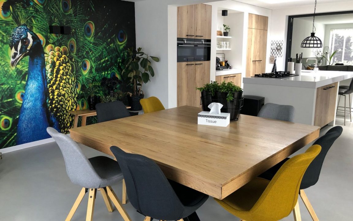 Eettafel met stoelen en open keuken op een naadloze microcement vloer, met pauwenbehang op de achterwand