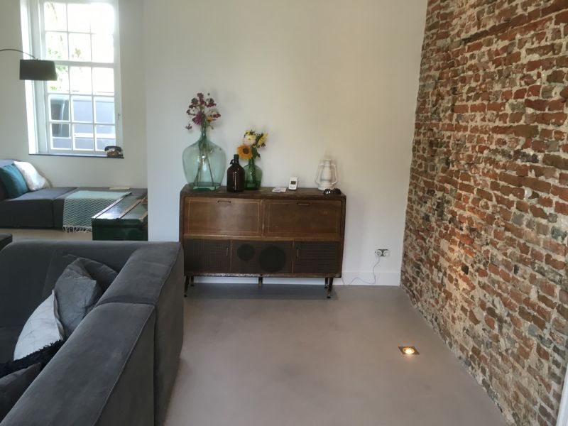 Industriële betonnen vloer in een zithoek in Son, met grijze bank, vintage dressoir en een wand van oude baksteen