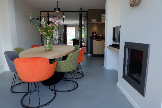 Eetkamer met ovale tafel en gekleurde stoelen bij een inbouwhaard, op een grijze microcement vloer